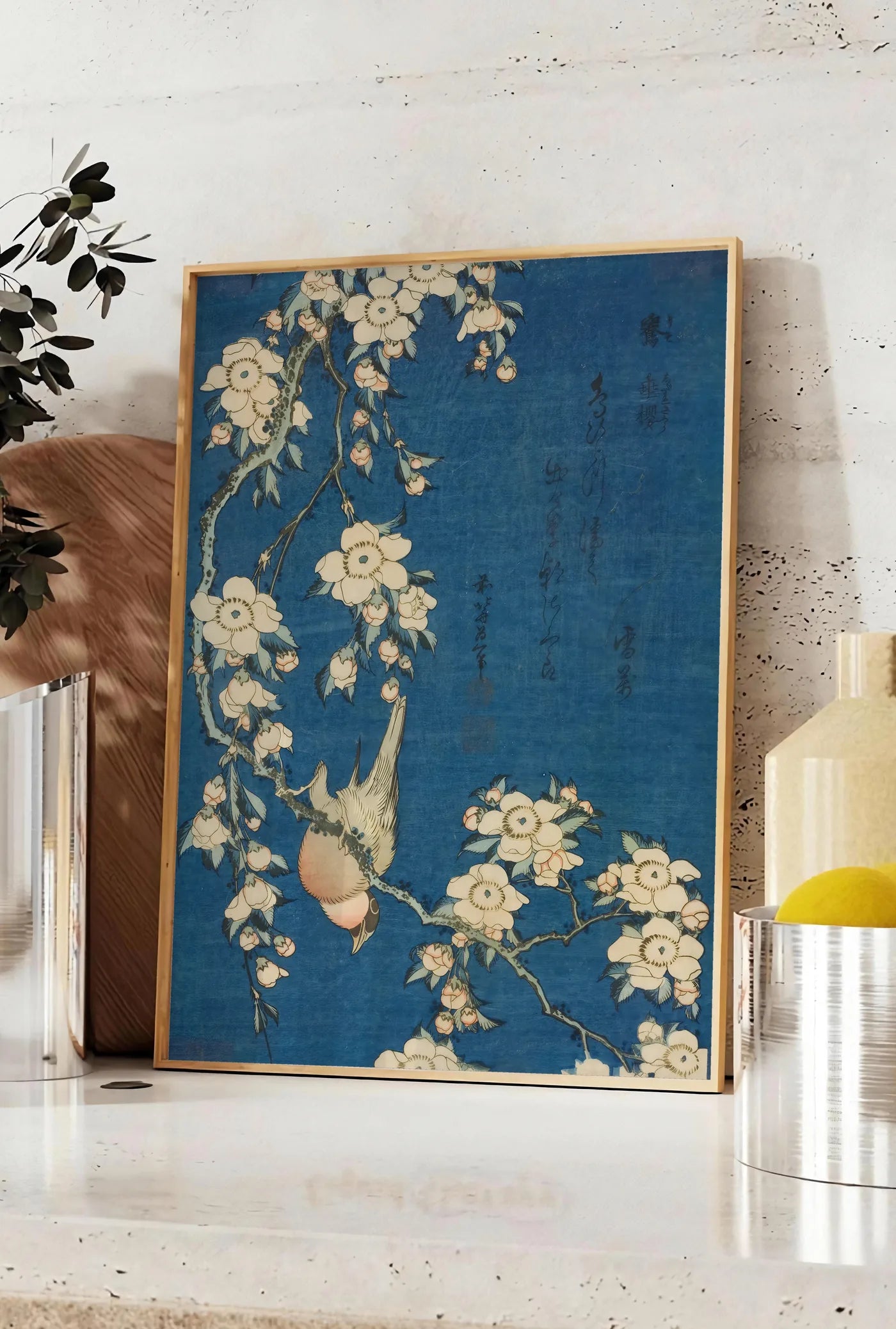 Affiche affiche Hokusai bouvreuil sur cerisier, posee sur un plan de travail de cuisine en pierre claire, entre une branche d'eucalyptus en vase et un bocal de citrons