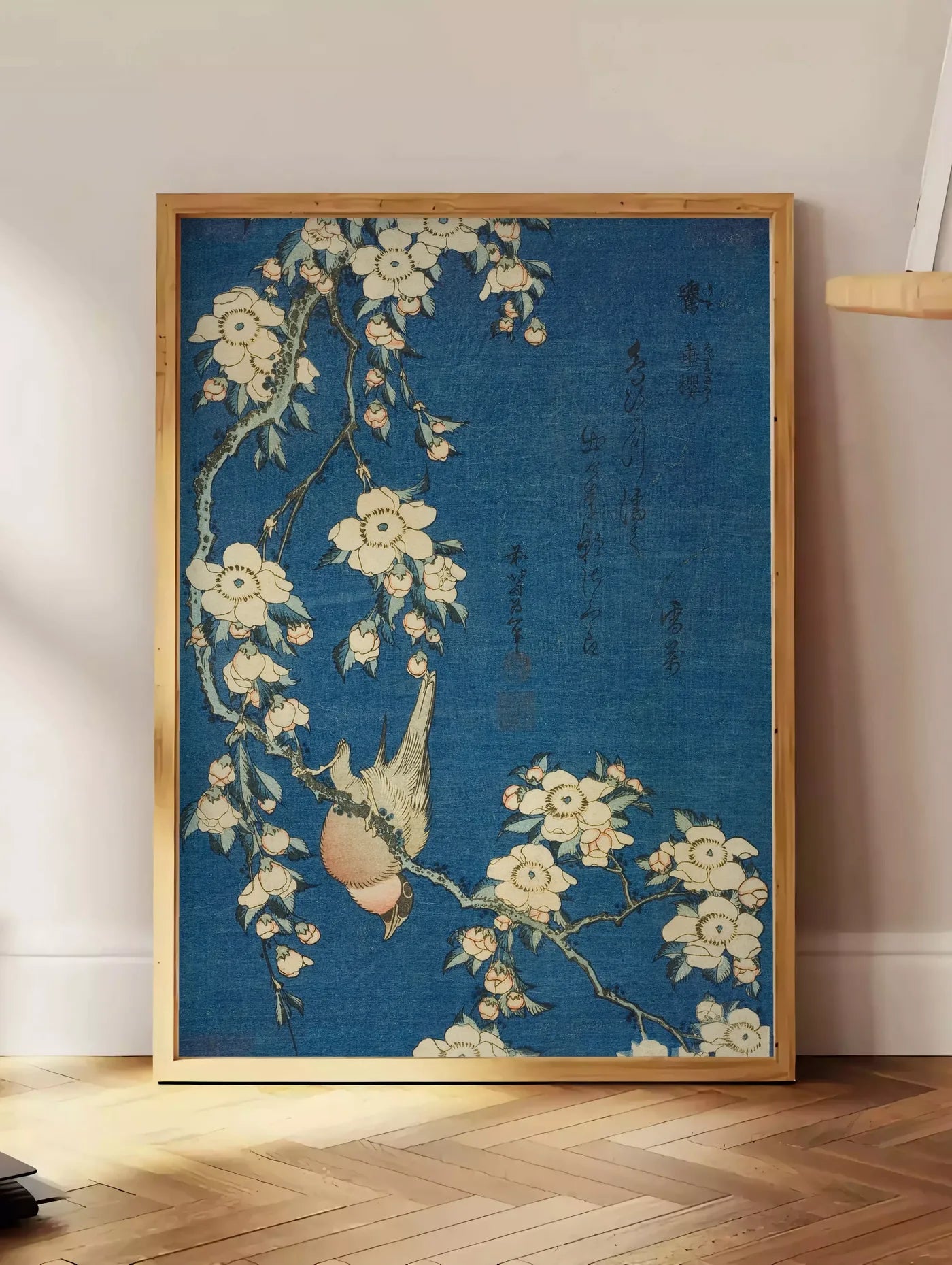 Estampe affiche Hokusai cerisier en fleurs et bouvreuil, posee au sol dans un coin salon, sur un parquet a chevrons, pres d'une table d'appoint en chene chargee de livres