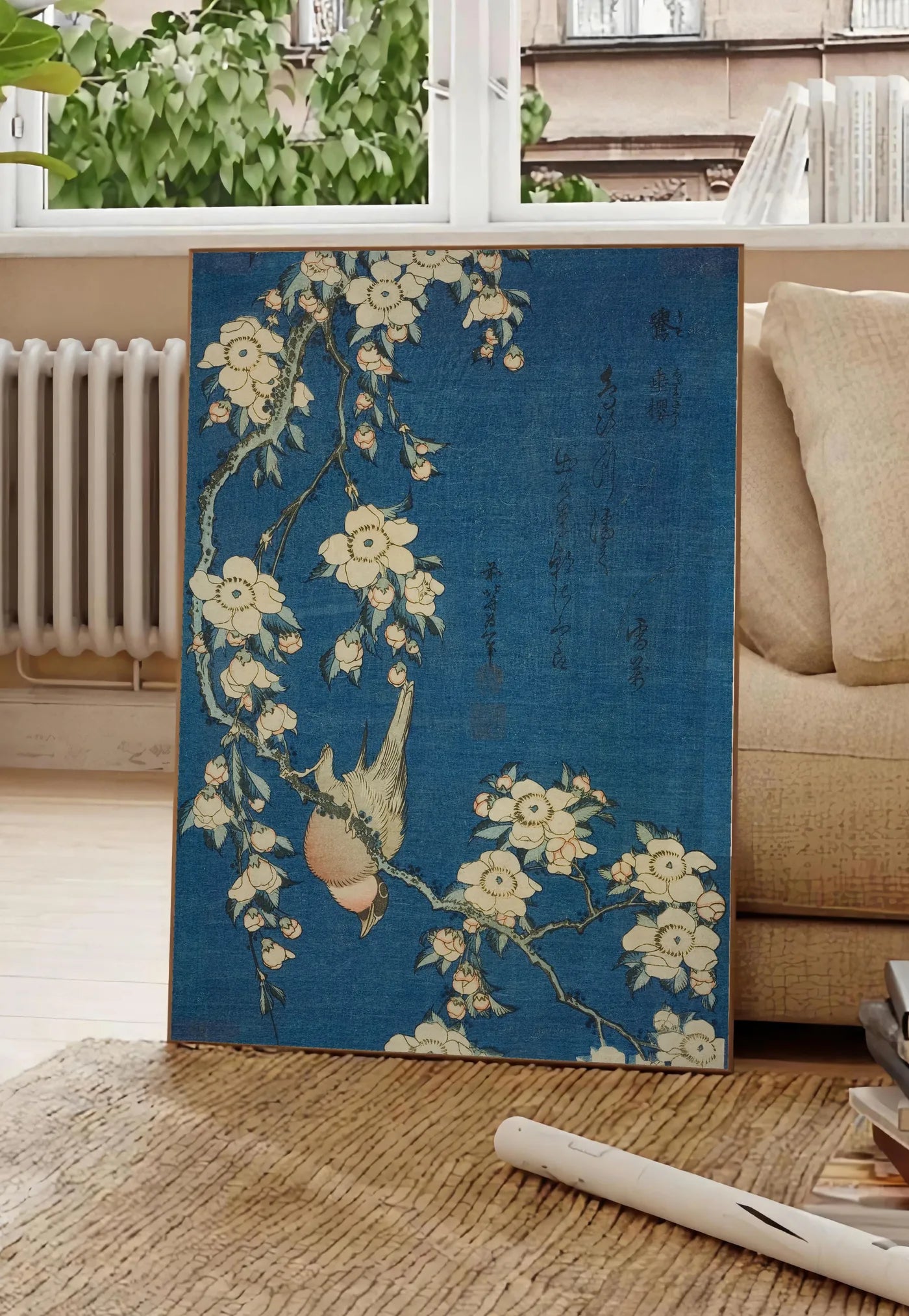 Poster affiche Hokusai cerisier en fleurs et bouvreuil, posee au sol dans un salon parisien, devant une grande fenetre donnant sur un immeuble haussmannien, entre un ficus et un canape creme