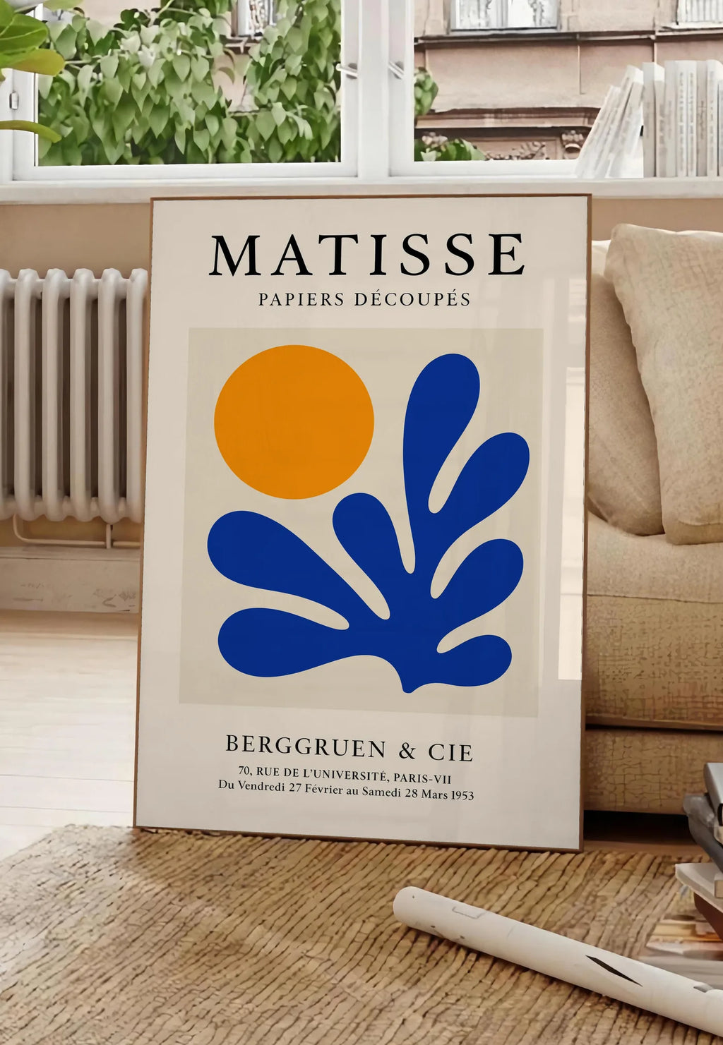 Poster corail Bleu et Soleil Orange en papiers decoupes Matisse sur fond colore, posee au sol dans un salon parisien, devant une grande fenetre donnant sur un immeuble haussmannien, entre un ficus et un canape creme